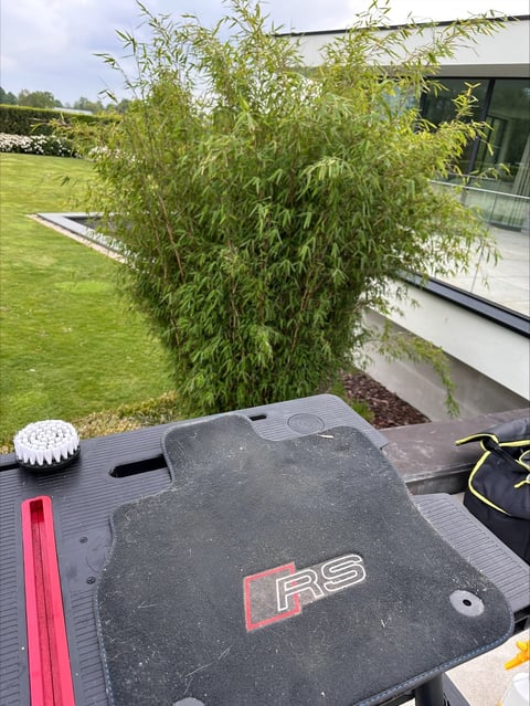 Black car floor mat with R9 logo on a table, with green garden, willow tree, and modern house visible in background